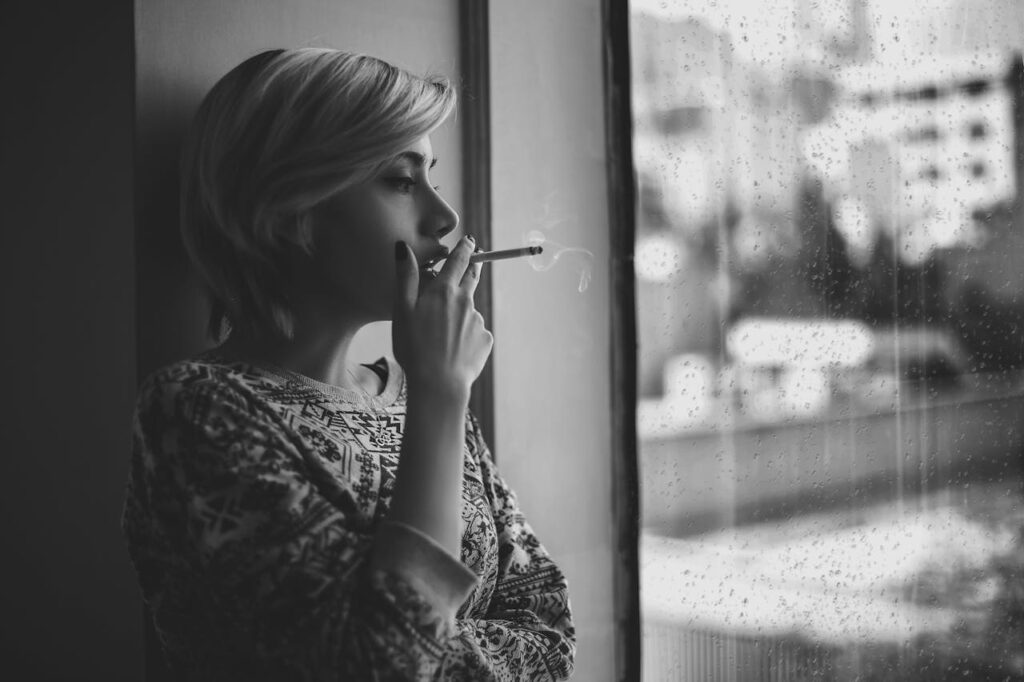 Black and white depressed female with short haircut smoking cigarette and looking out window at rainy city